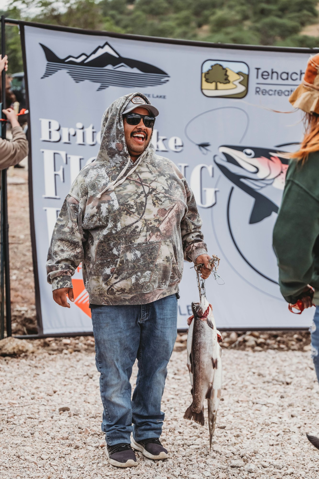 A person in a camo hoodie holds a fish, smiling at a fishing event with a sign in the background.