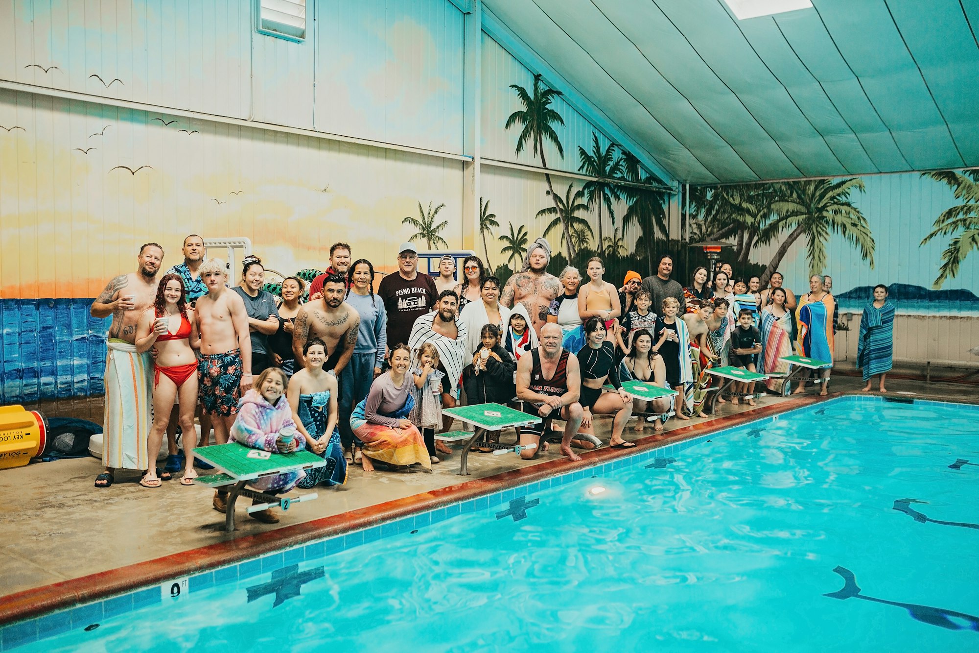 A group of people posing by an indoor pool with a tropical mural backdrop.