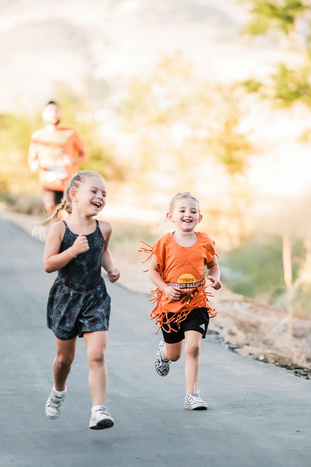 Two young girls joyfully running on a paved path, with a person in the background also running.