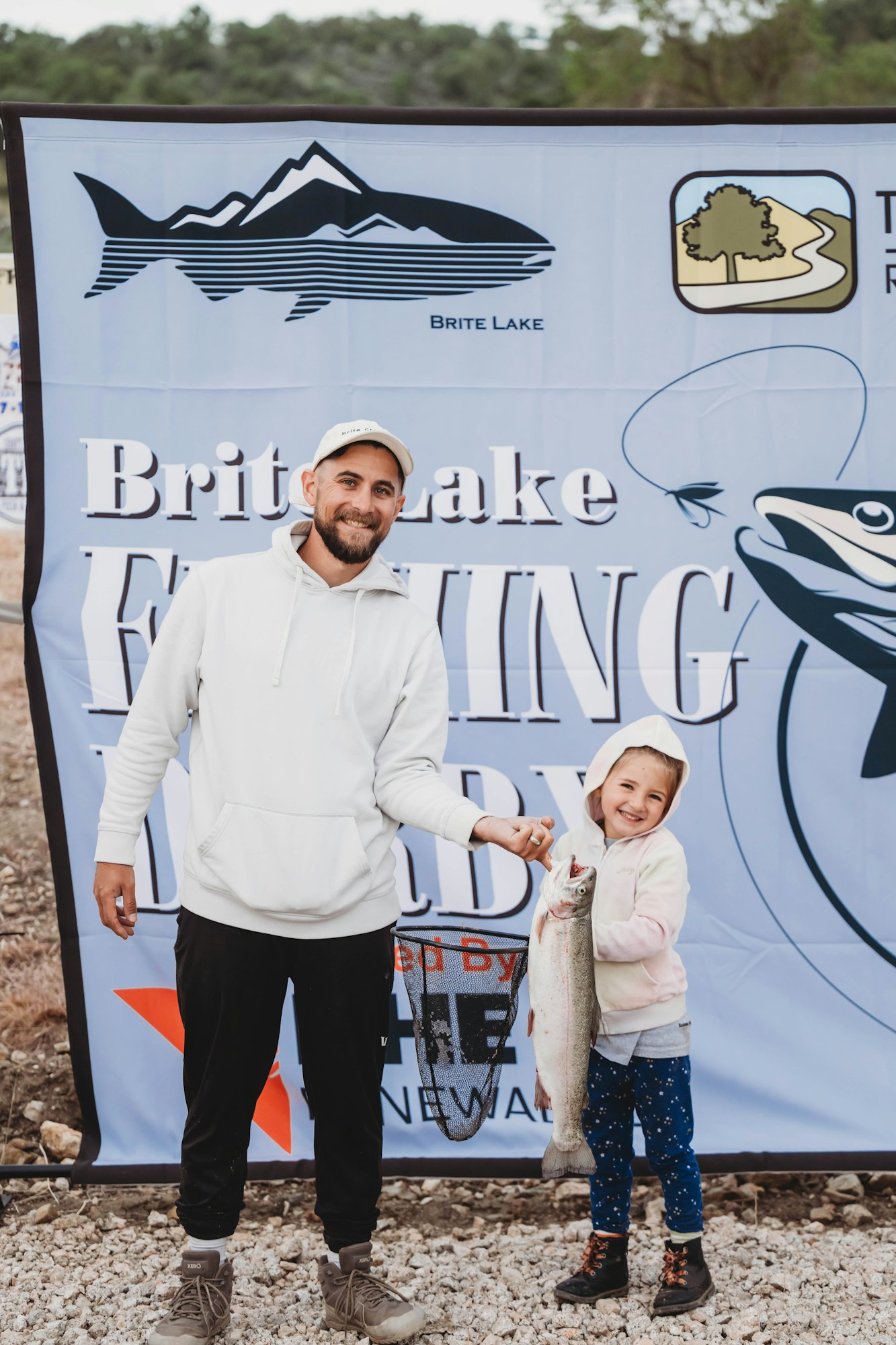 A person and child smile, holding a fish at the Brite Lake Fishing Derby, standing in front of an event banner.