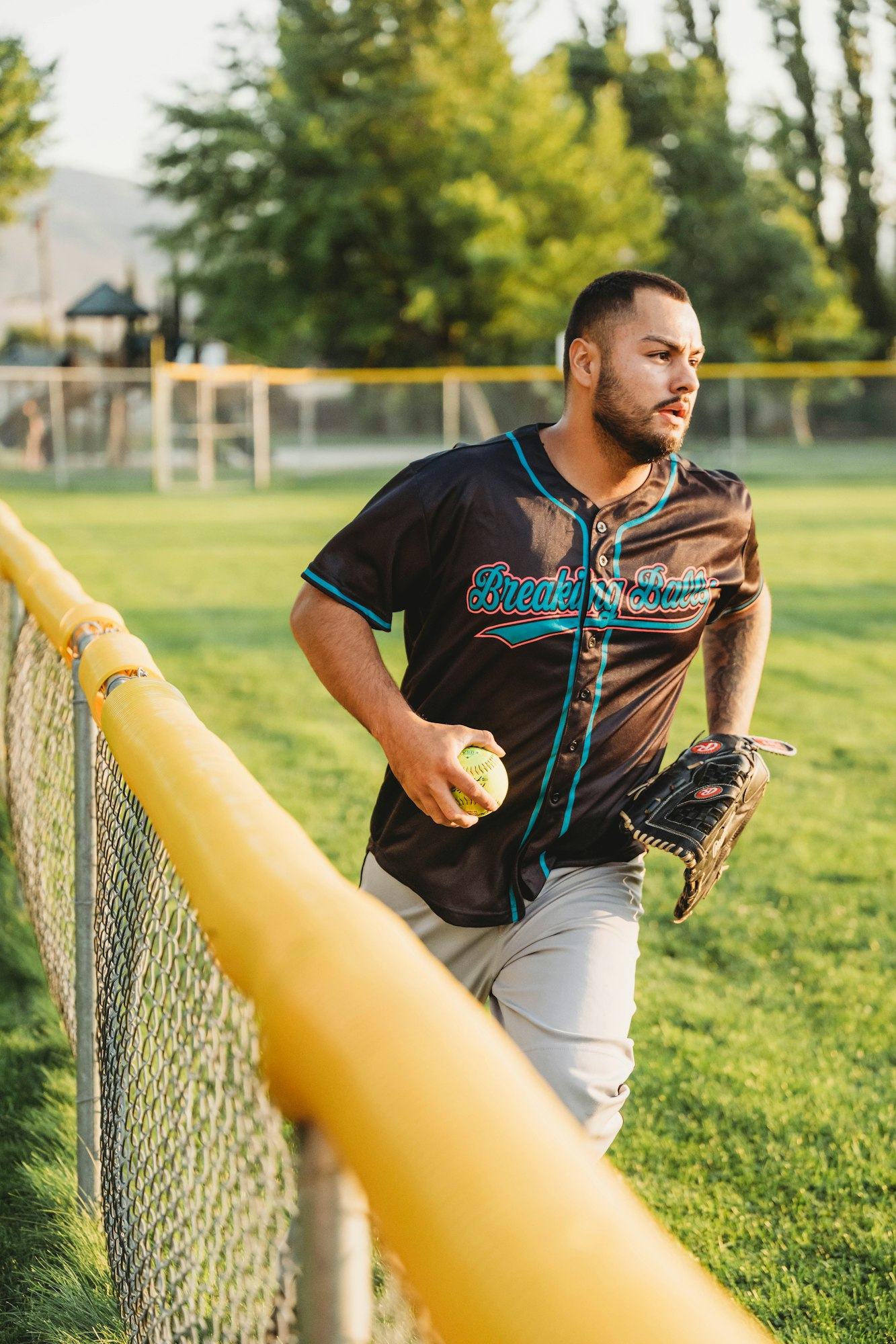 A man in a baseball uniform with a glove and ball, leaning on a fence at a park.