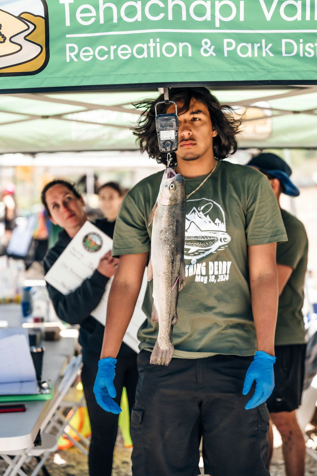 A person weighing a fish at a fishing derby, with an observer holding a certificate in the background.
