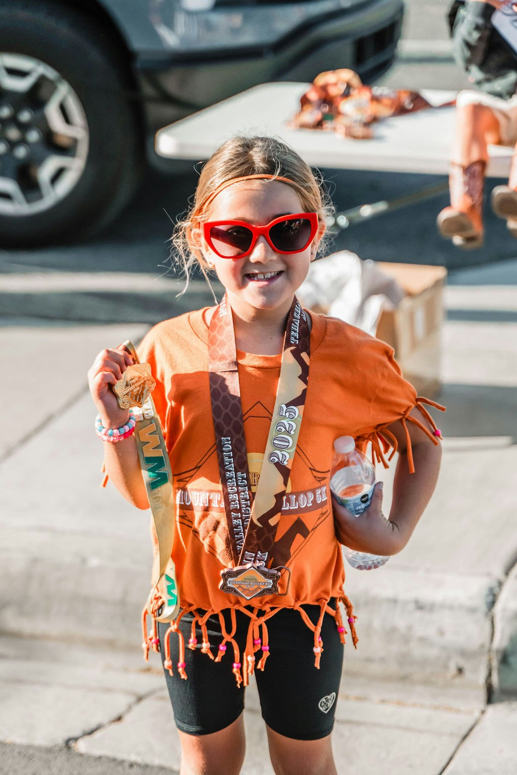 A smiling child in sunglasses holds medals and a water bottle, wearing an orange shirt, standing outdoors near a parked car.