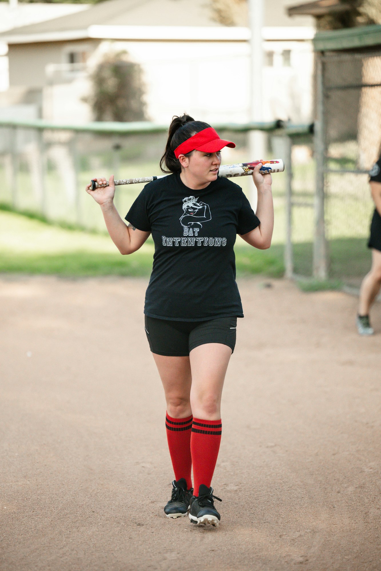 A woman in a black t-shirt and red cap stands on a dirt field, holding a softball bat over her shoulder.