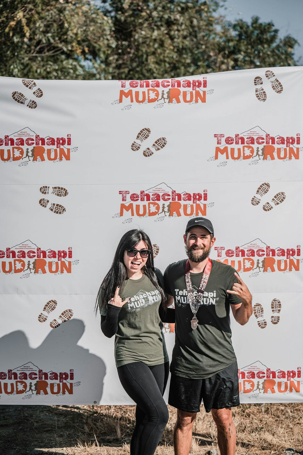 Two people posing in front of a "Tehachapi Mud Run" backdrop, wearing matching event shirts and displaying medals.