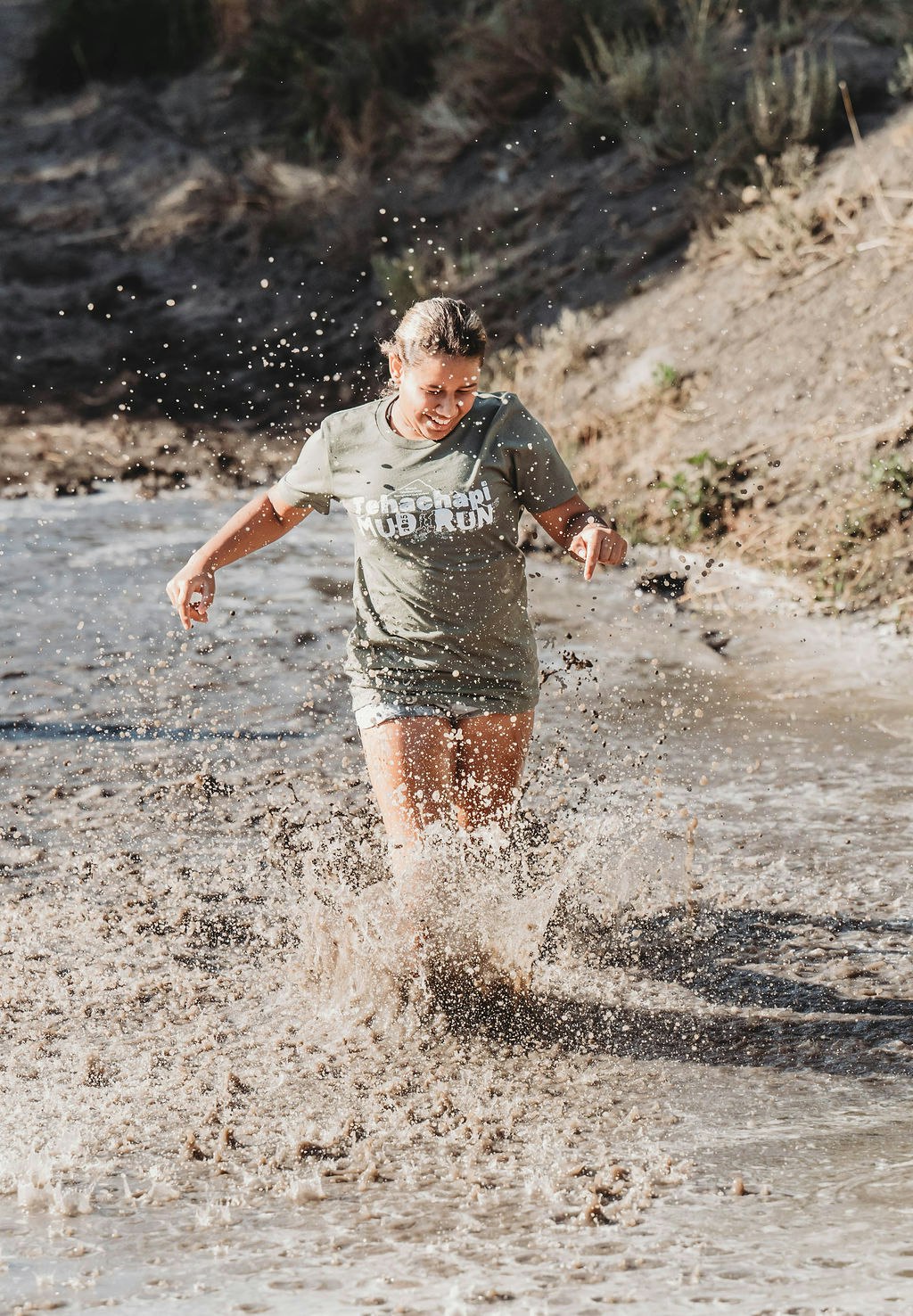 Person running through muddy water with "Tehachapi Mud Run" on their shirt, splashing around.