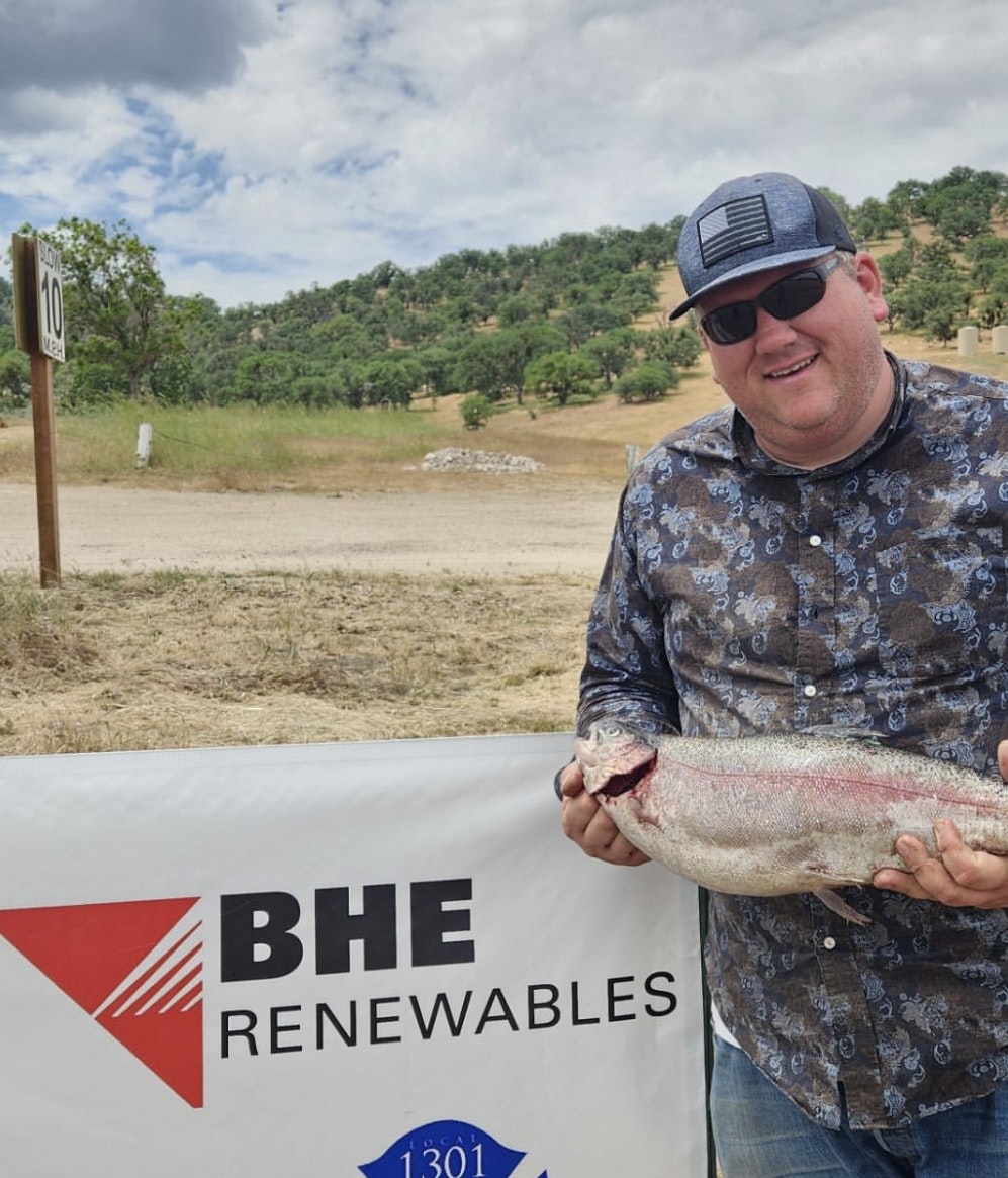 A man holding a fish in front of a "BHE Renewables" banner, outdoors with trees in the background.