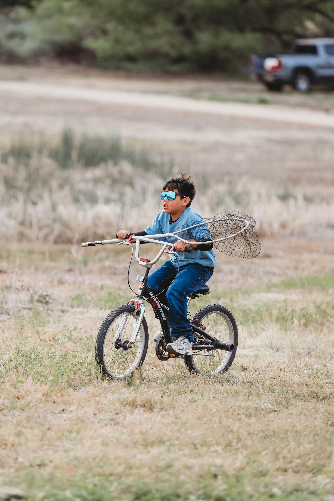 A kid in blue sunglasses rides a bike across a grassy field, holding a fishing net.