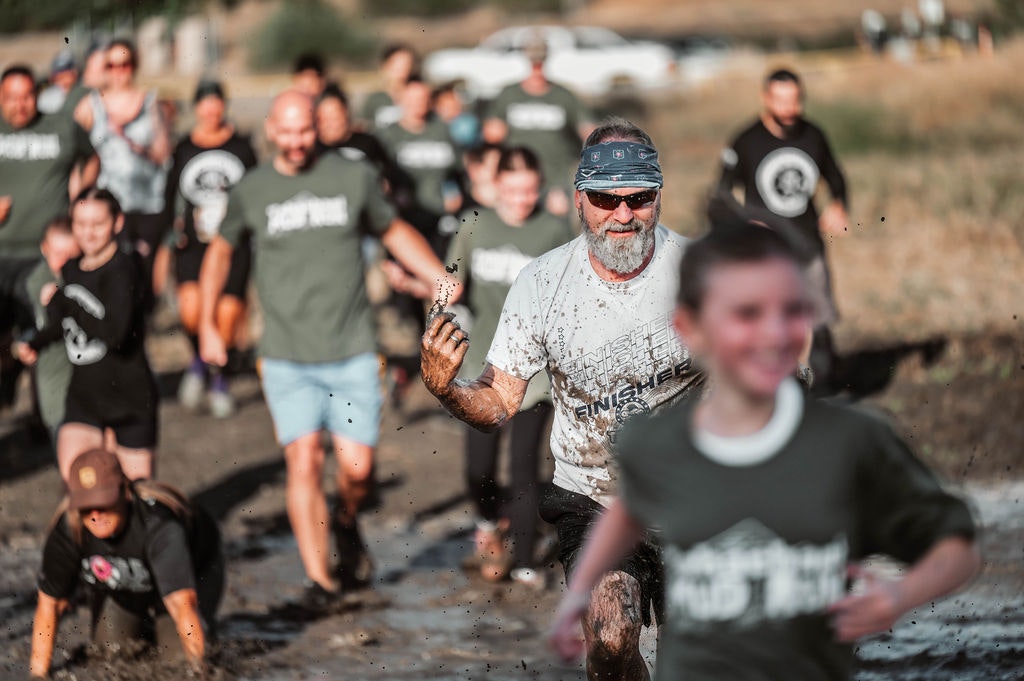 Adults and children run through a muddy obstacle course, smiling and enjoying the activity.