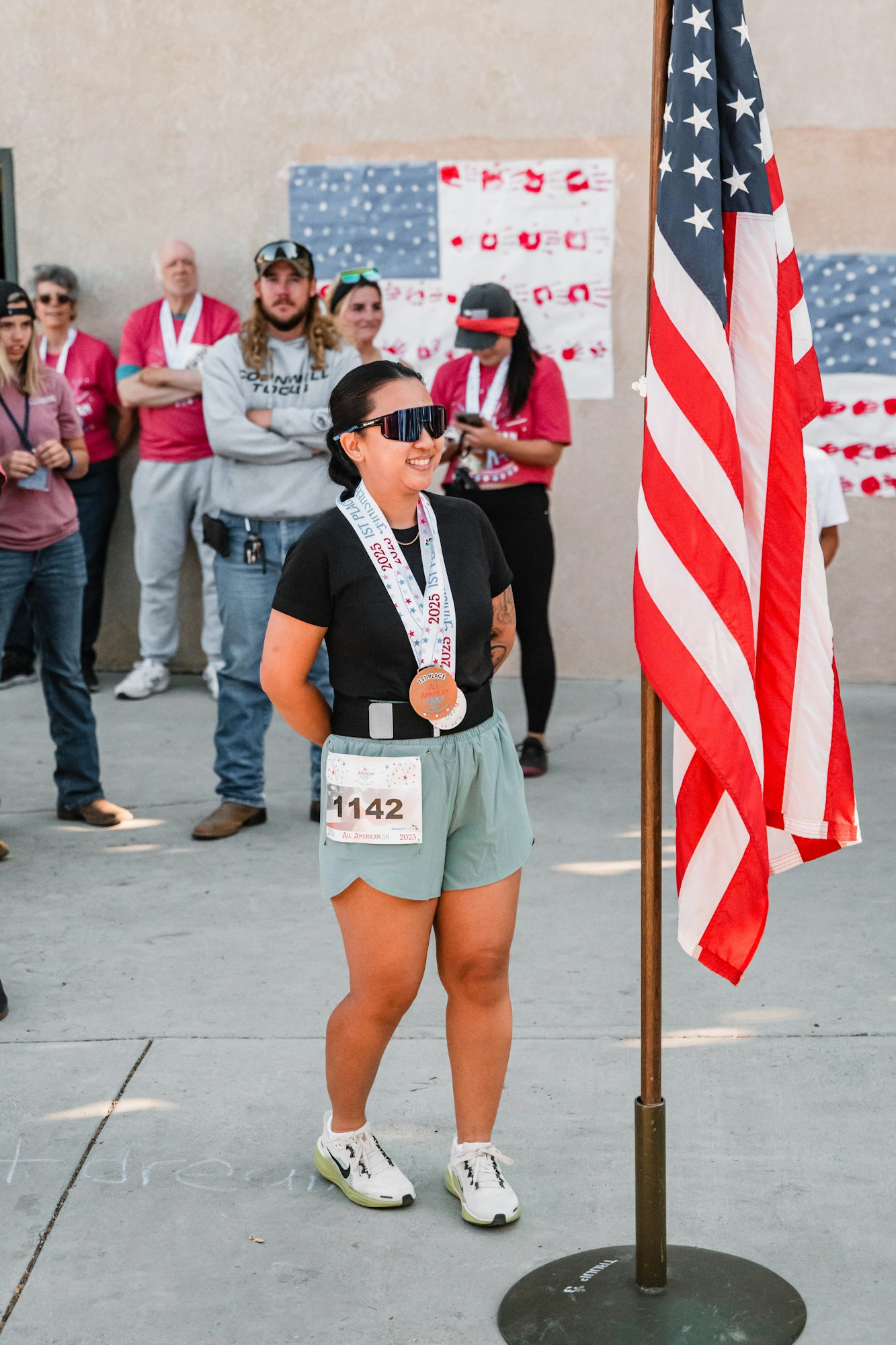 A person with a medal stands smiling near a U.S. flag, surrounded by a group wearing medals and event numbers.