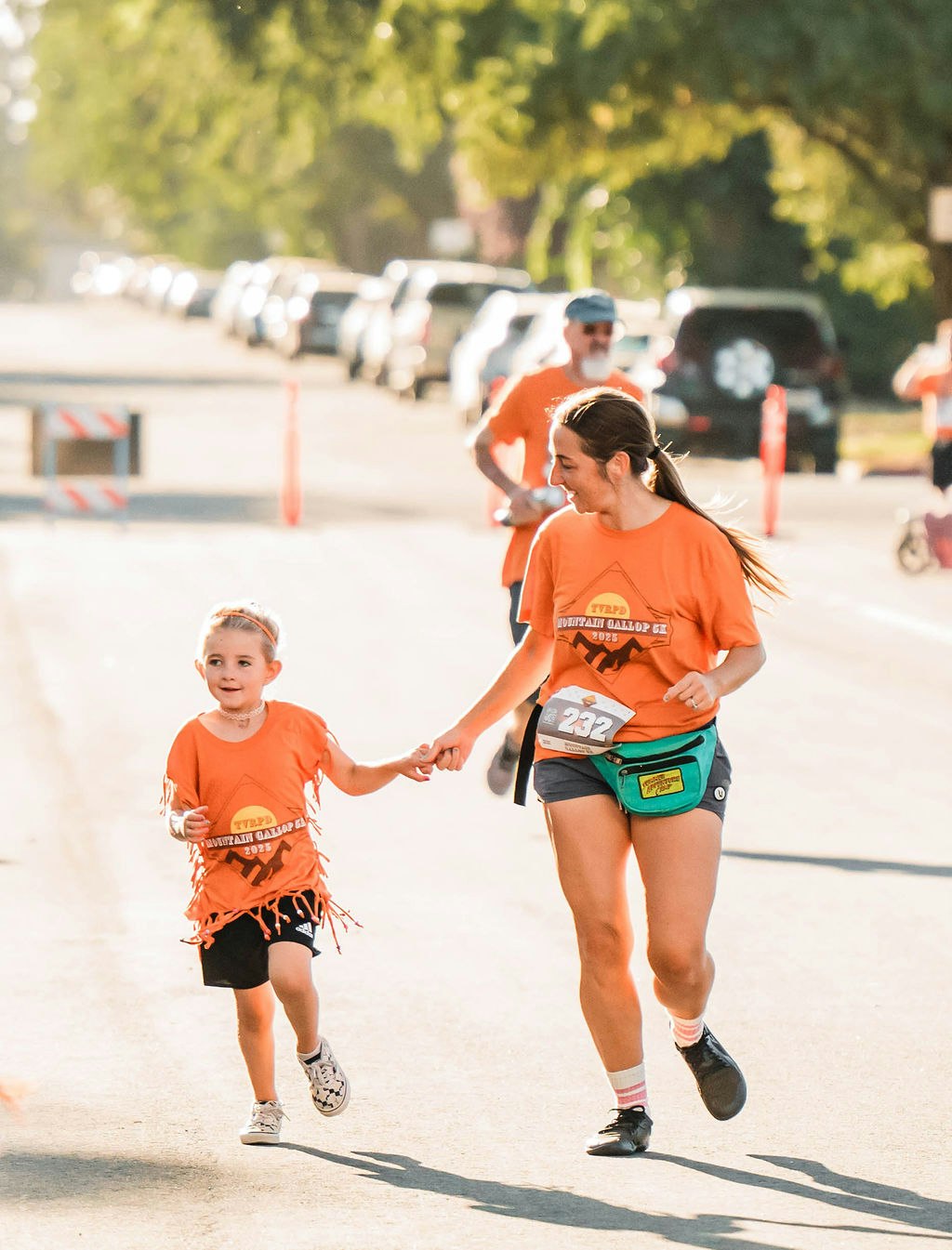 Two people in orange shirts running hand in hand on a sunny street during a race event, with other runners and parked cars in the background.