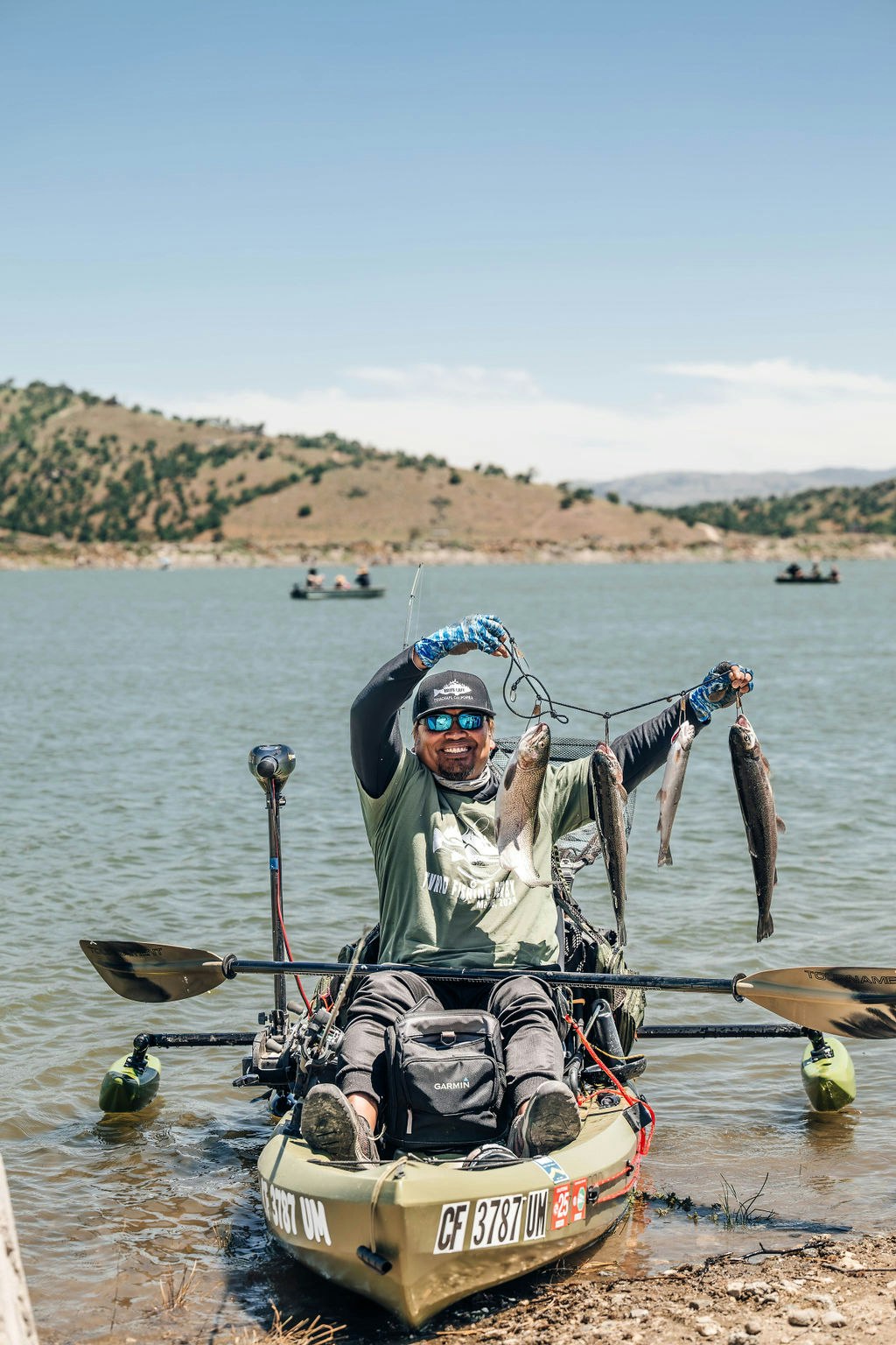 A man in a kayak holding fish with a lake and hills in the background.