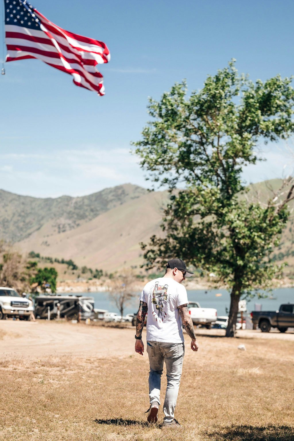A person walking outdoors with US flag fluttering in the background, green trees, and a mountainous landscape.