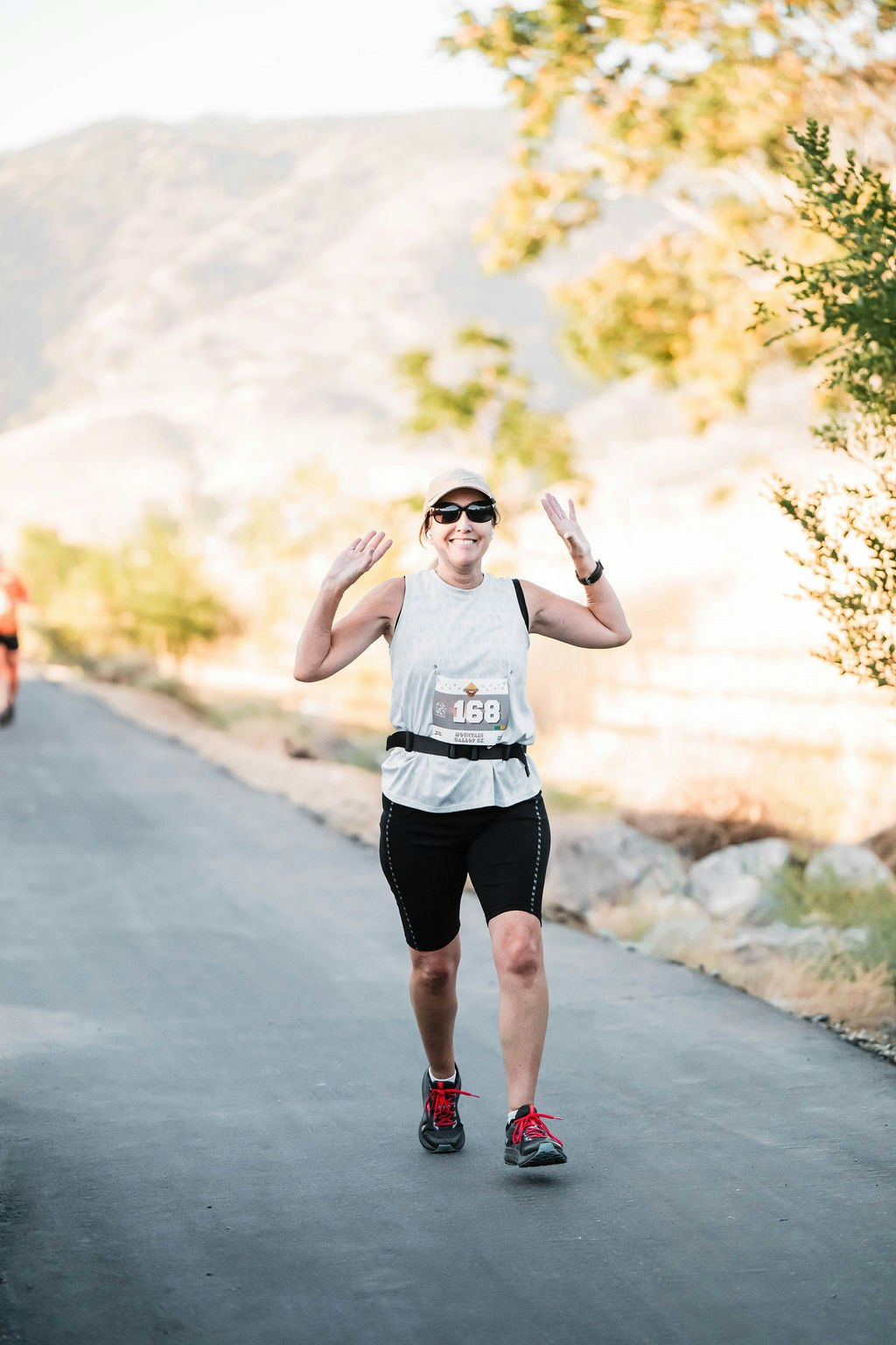 A woman wearing a race bib and athletic gear runs on a paved path, smiling with arms raised, against a blurred outdoor background.