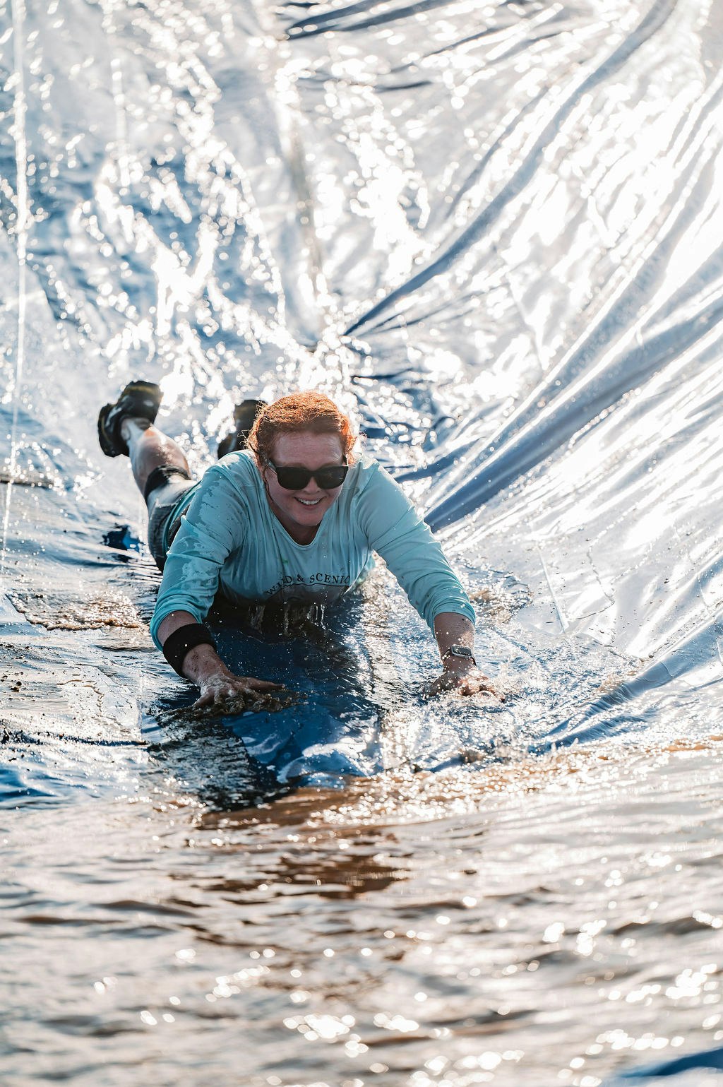 Person sliding down a water slide, smiling, wearing sunglasses and a light blue shirt.