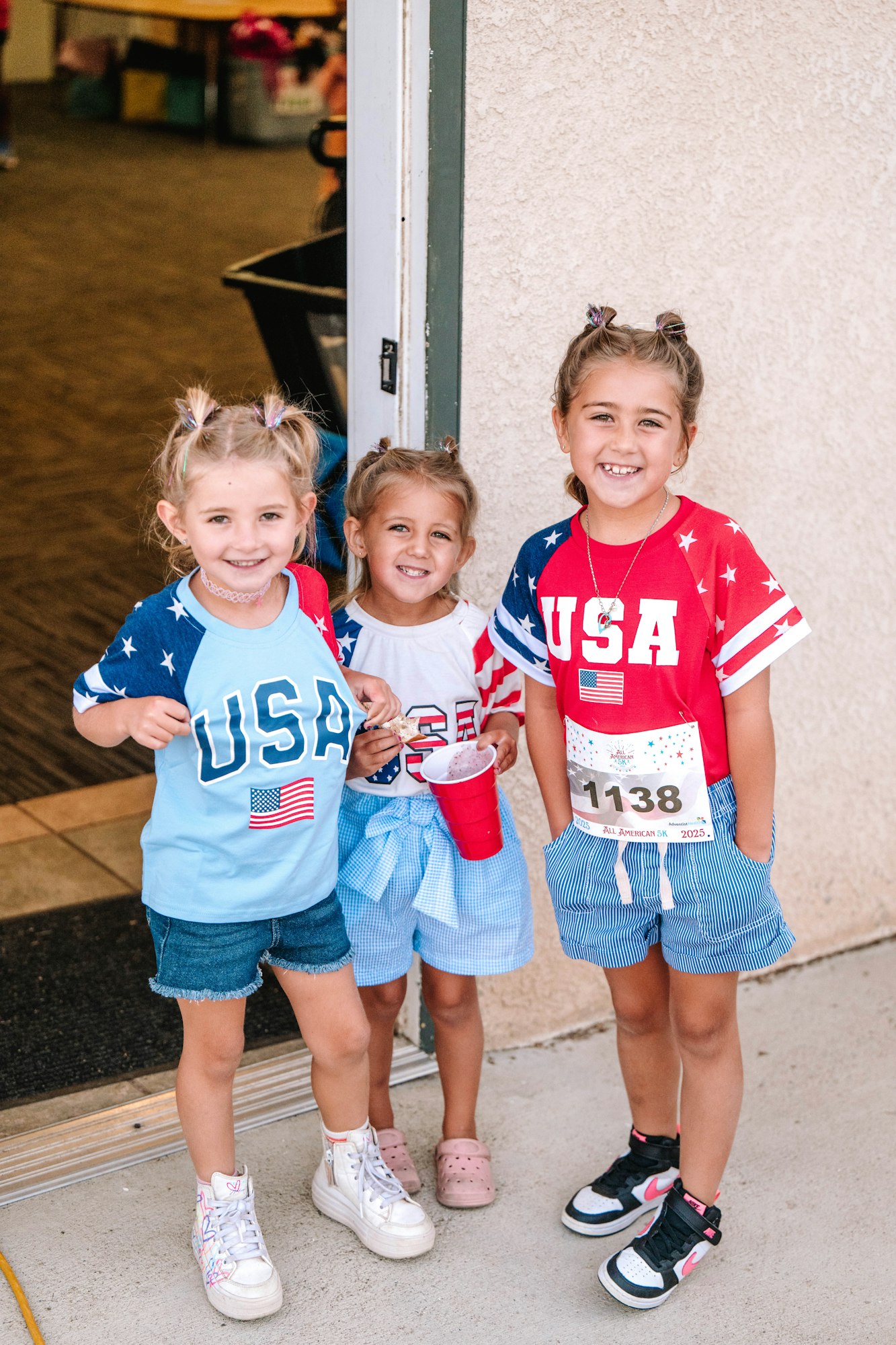 Three kids wearing USA-themed outfits, smiling, standing by a door.