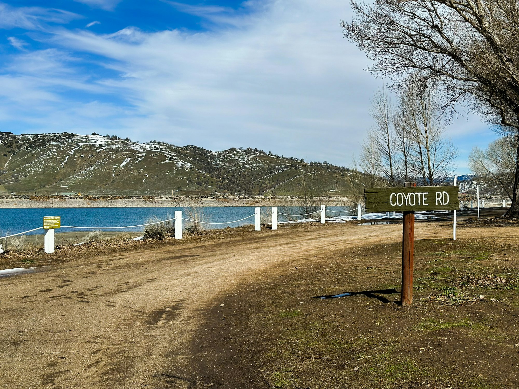 A "COYOTE RD" sign, dirt road, fence, lake, and hills with sparse snow under a blue sky with clouds.