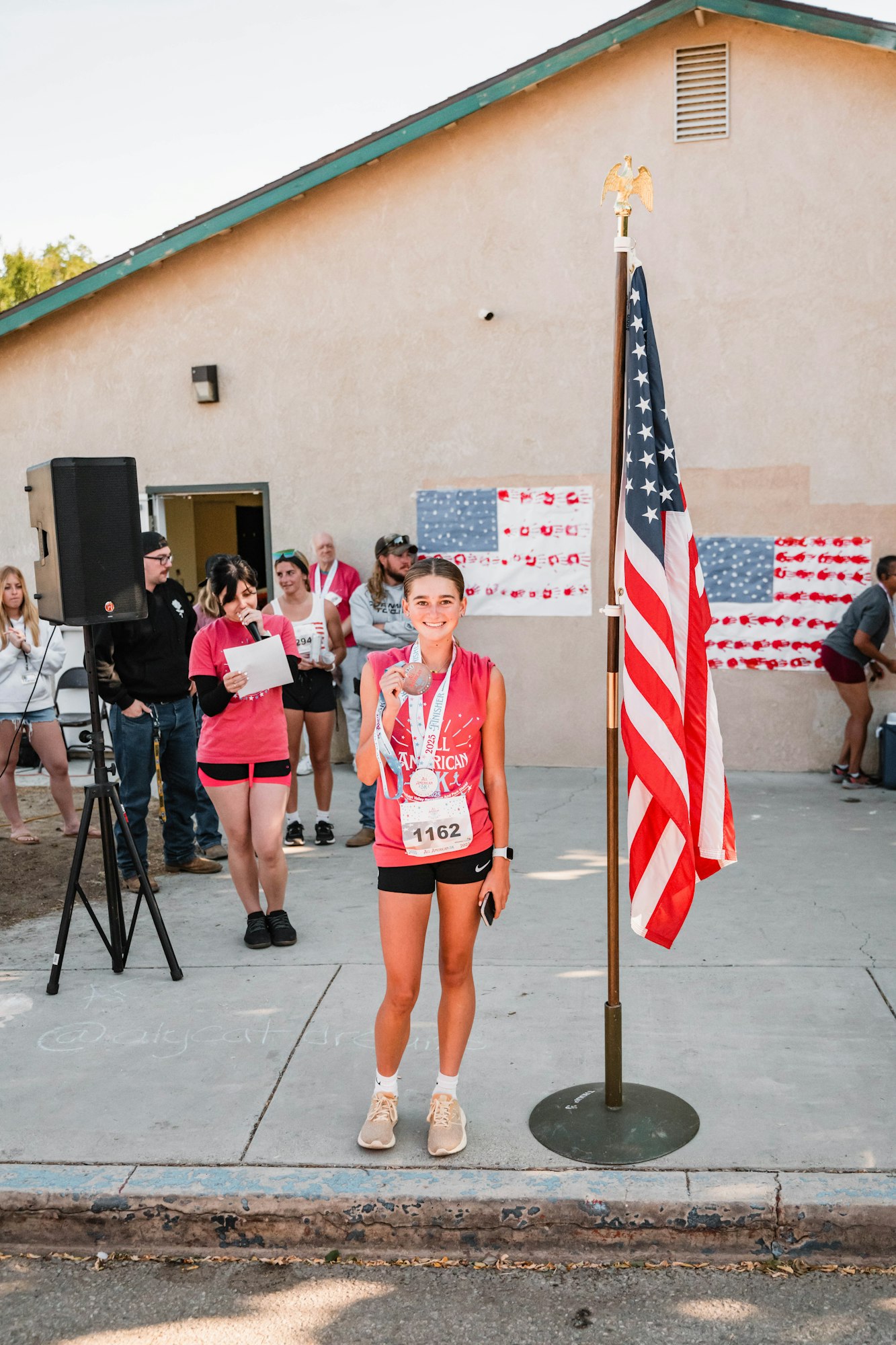 A person wearing a medal stands beside an American flag. Others are gathered in the background outside a building.