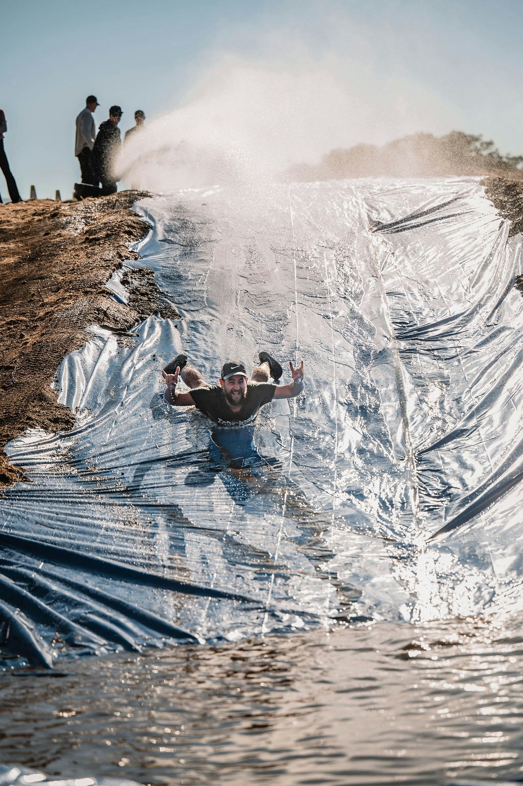 Man sliding down a plastic water slide, making hand gestures, smiling. Sunlit scene with onlookers and water splashes.