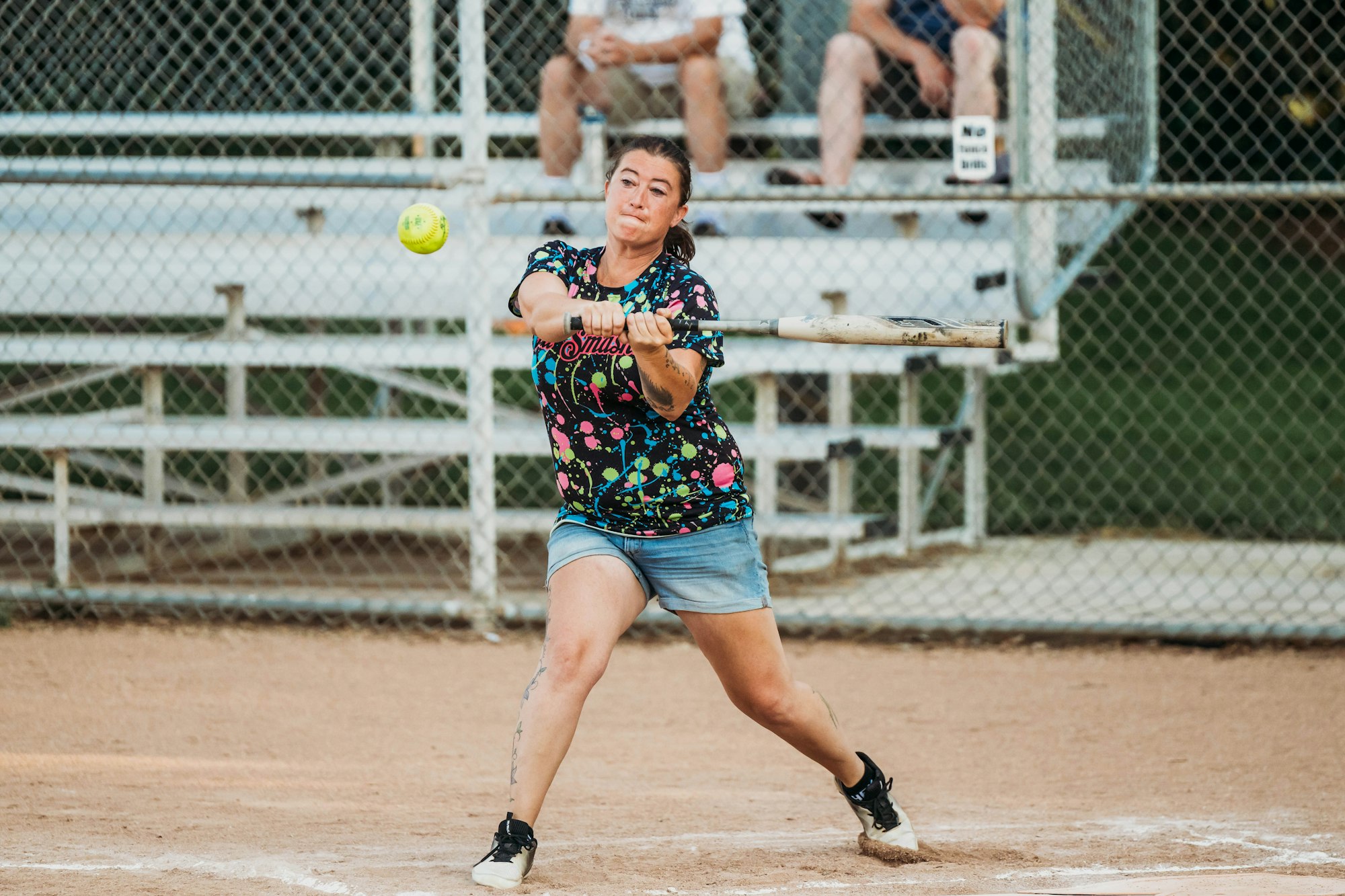 A person swinging a bat at a softball on a dirt field with spectators in the background.