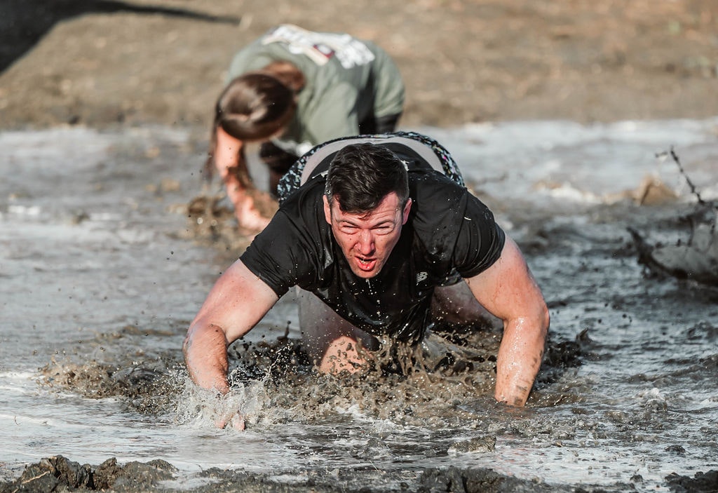 Two people crawling through a muddy obstacle course, determined expressions, splashing water.