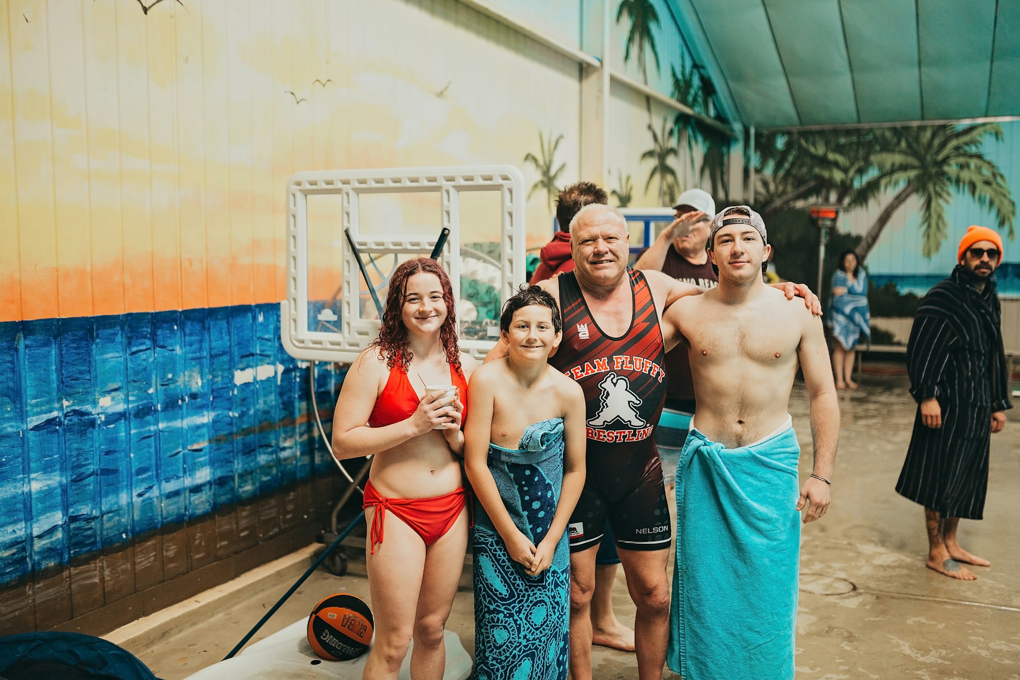 A group of people in swimwear smiling, with a basketball and tropical mural in the background.