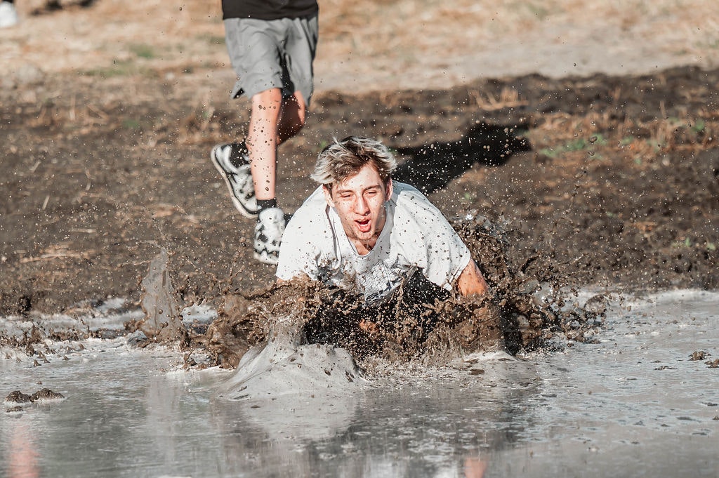 A person sliding face-first into a muddy puddle with splashes around them. Another person runs behind.