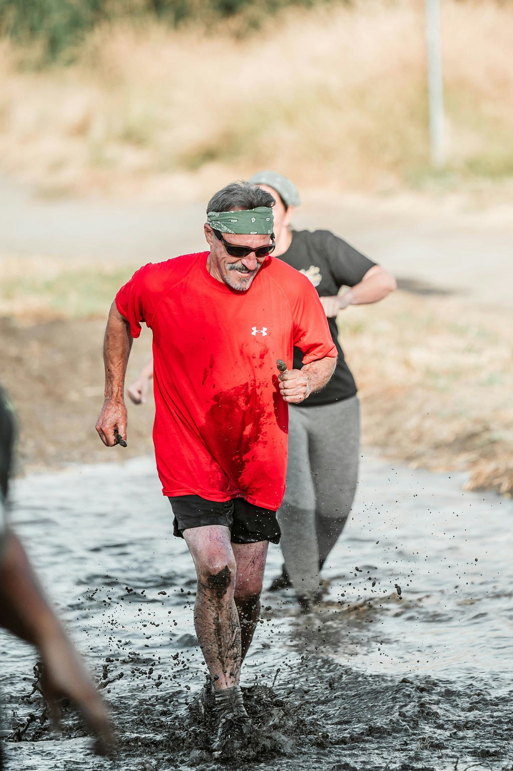 A man in a red shirt and bandana runs through mud, surrounded by others.