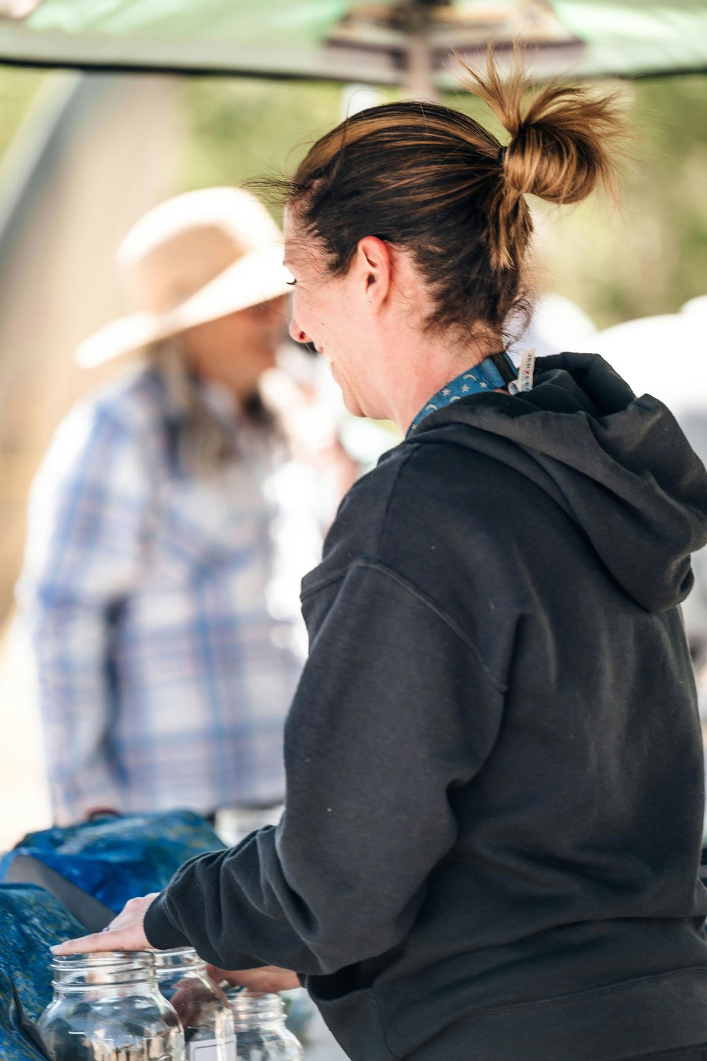 A person with a ponytail, wearing a hoodie, and holding a jar, with another person in a hat blurred in the background.