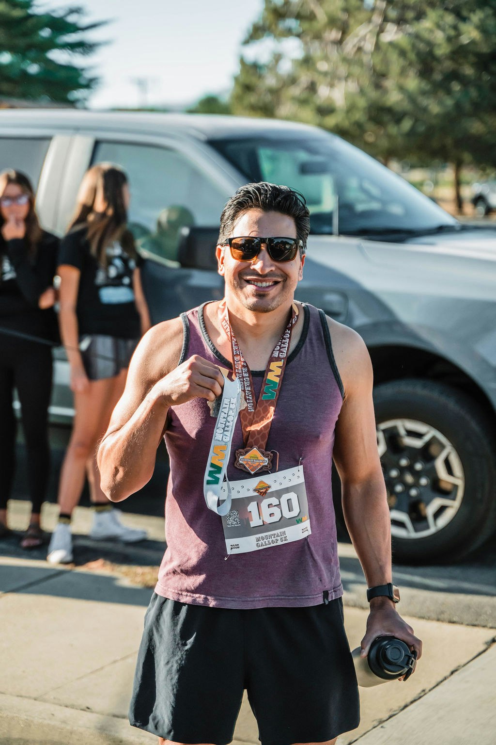 Smiling man wearing a race bib and medal, holding a bottle, standing in front of a van.