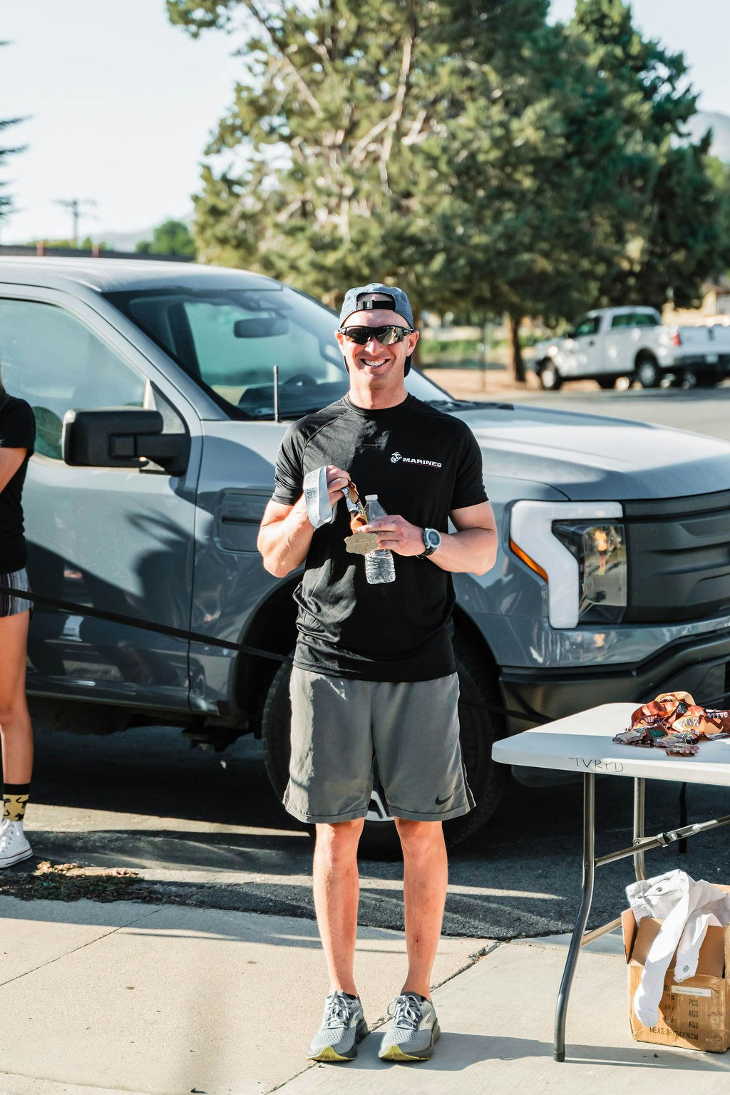 A person in athletic wear holding a drink, standing by a table with medals, next to a parked truck, outdoors.