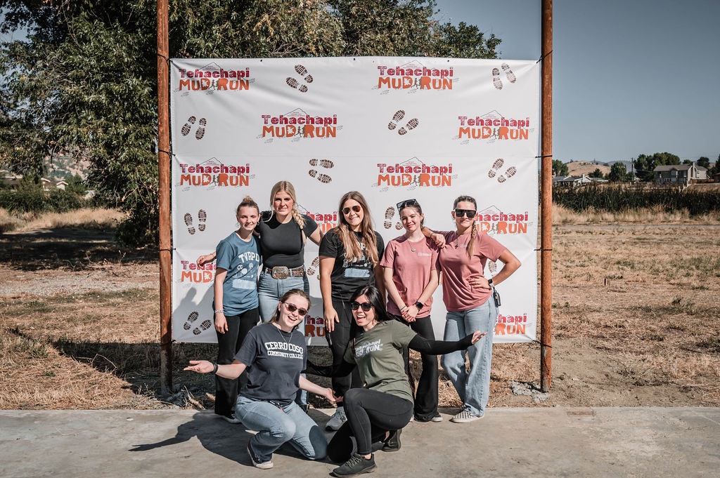 A group of seven people poses in front of a "Tehachapi Mud Run" banner outdoors.