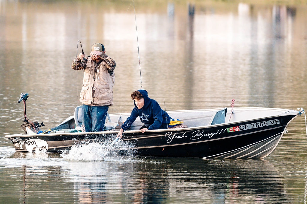 Two people on a small boat, one fishing and the other operating an outboard motor, on calm water.