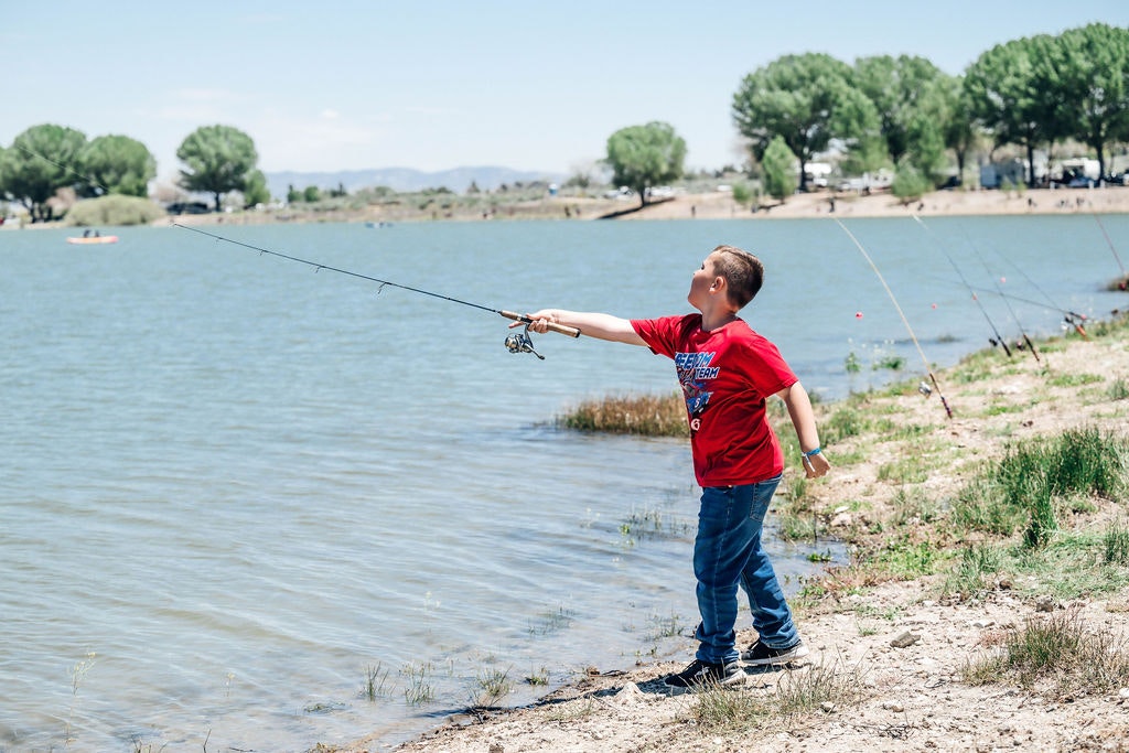 A child is fishing by a lakeside on a sunny day, with trees and people in the background.