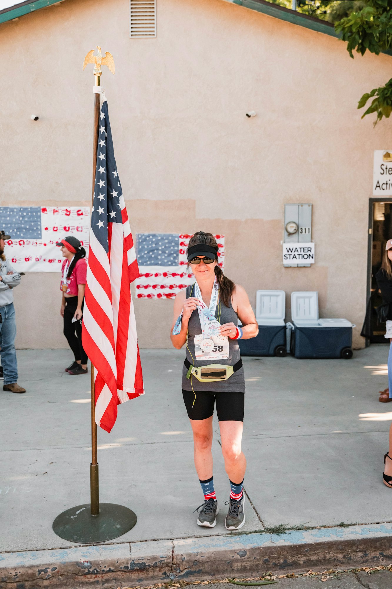 Runner with a medal stands beside a US flag, wearing sporty attire. Background shows a water station and people.