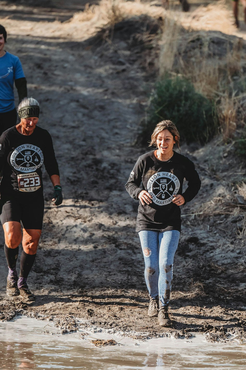 Two people in "Lion Heart" shirts are running through a muddy area in an outdoor event.