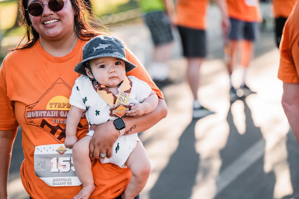 A smiling person holds a baby wearing a hat and a medal. They're at an event, with others in orange shirts in the background.