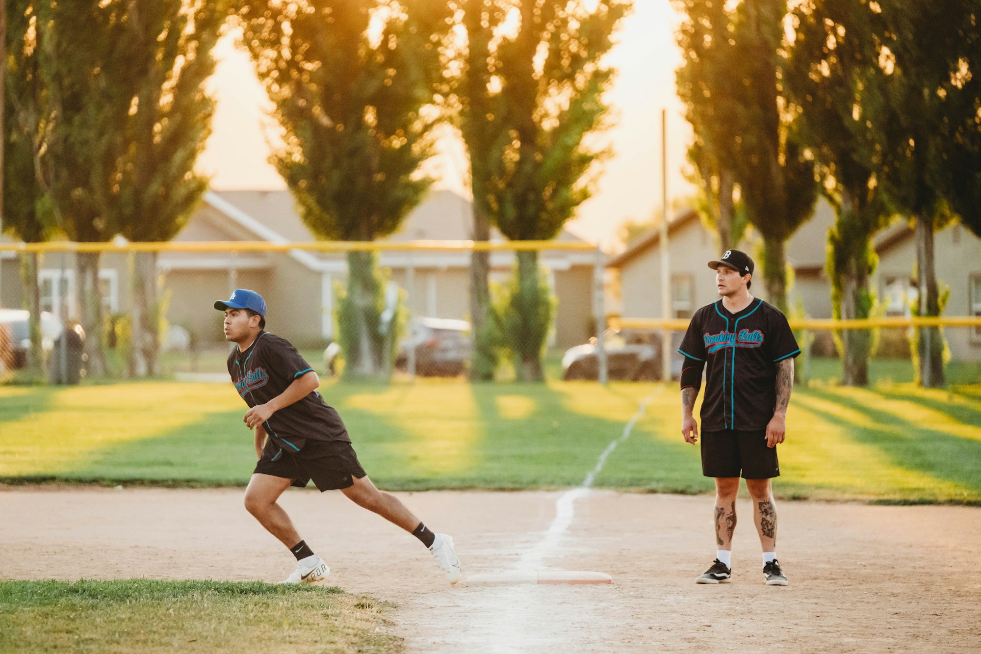Two players in baseball uniforms on a dusty field, one running bases and the other watching. Sunset and trees in the background.