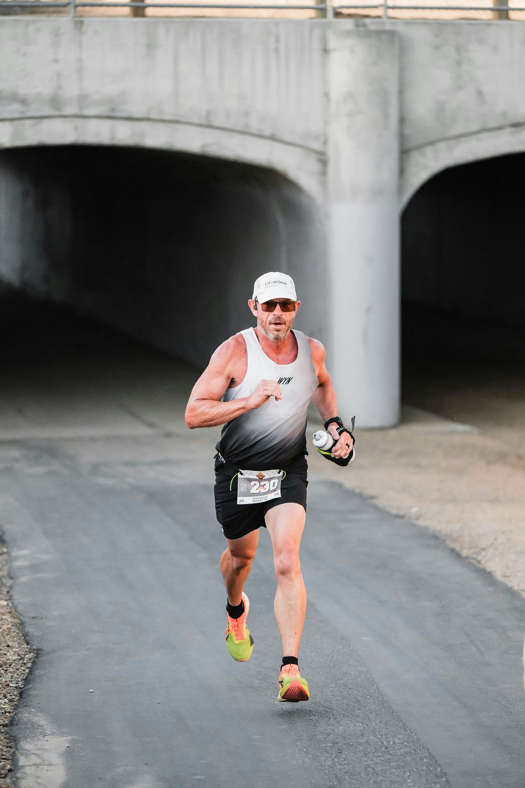 A person running on a paved path, wearing a hat, sunglasses, and numbered bib, holding a water bottle.