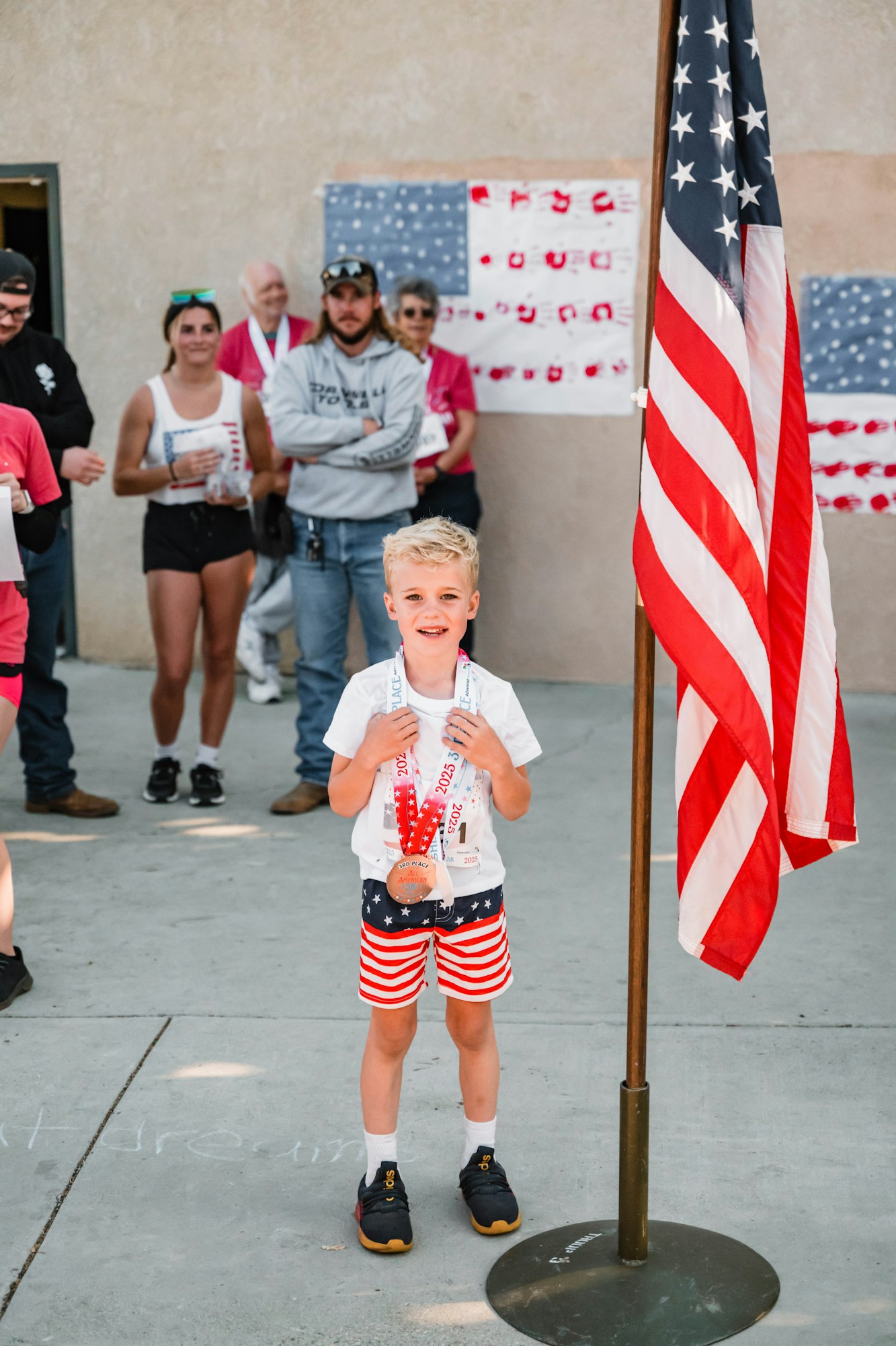A young boy in patriotic attire holds medals, standing beside the American flag. People are gathered and watching in the background.