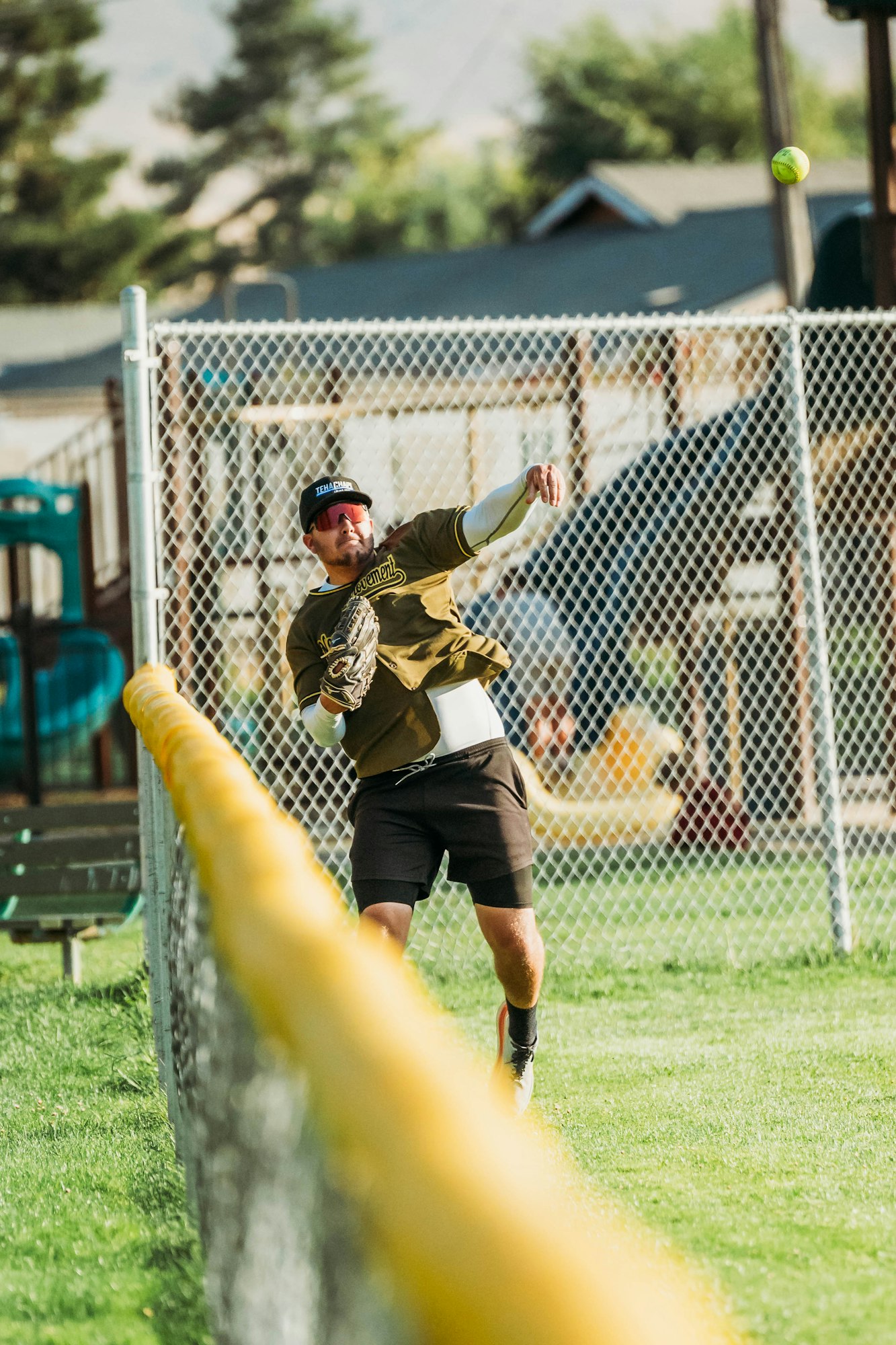 A person throws a softball, wearing a cap, sunglasses, and sports attire, near a chain-link fence.