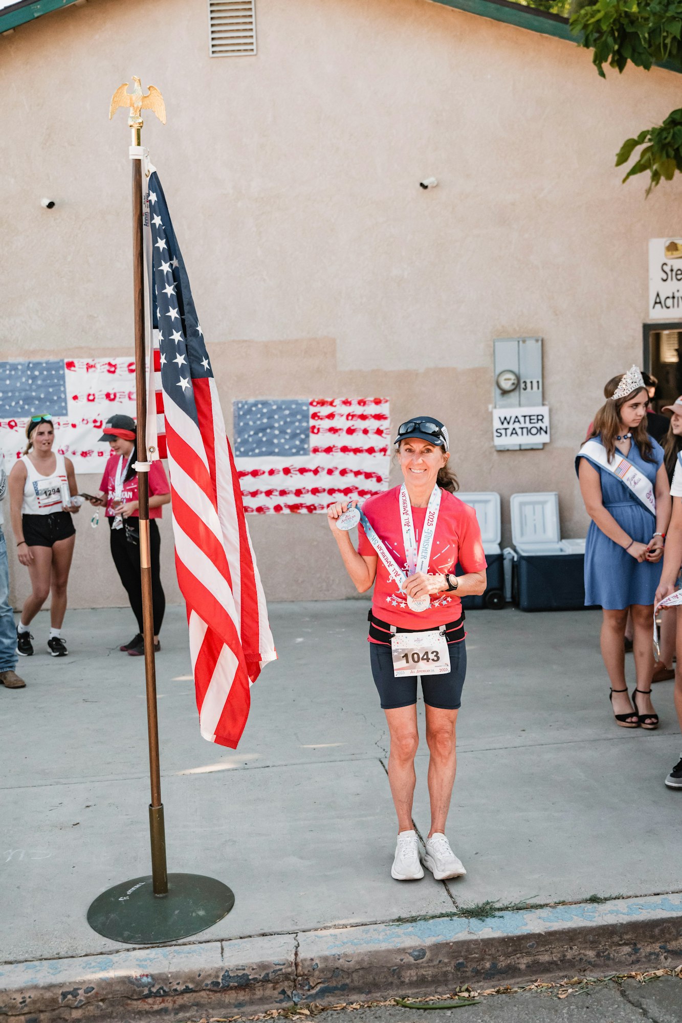 A runner with bib 1043 holds medals beside a U.S. flag. Others stand nearby. Decorated wall and "water station" sign in the background.