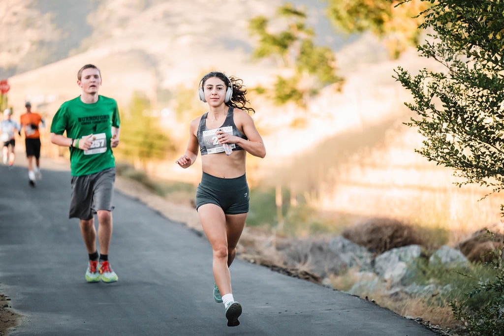 Two people running on a paved road with others in the background. Sunny day with trees and hills around.