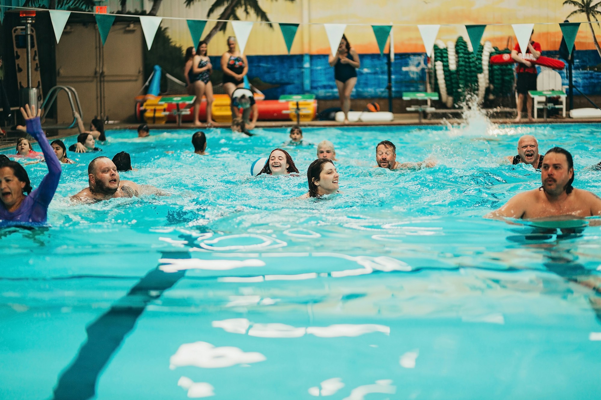 People enjoying a swim in an indoor pool with a mural background and spectators on the side.