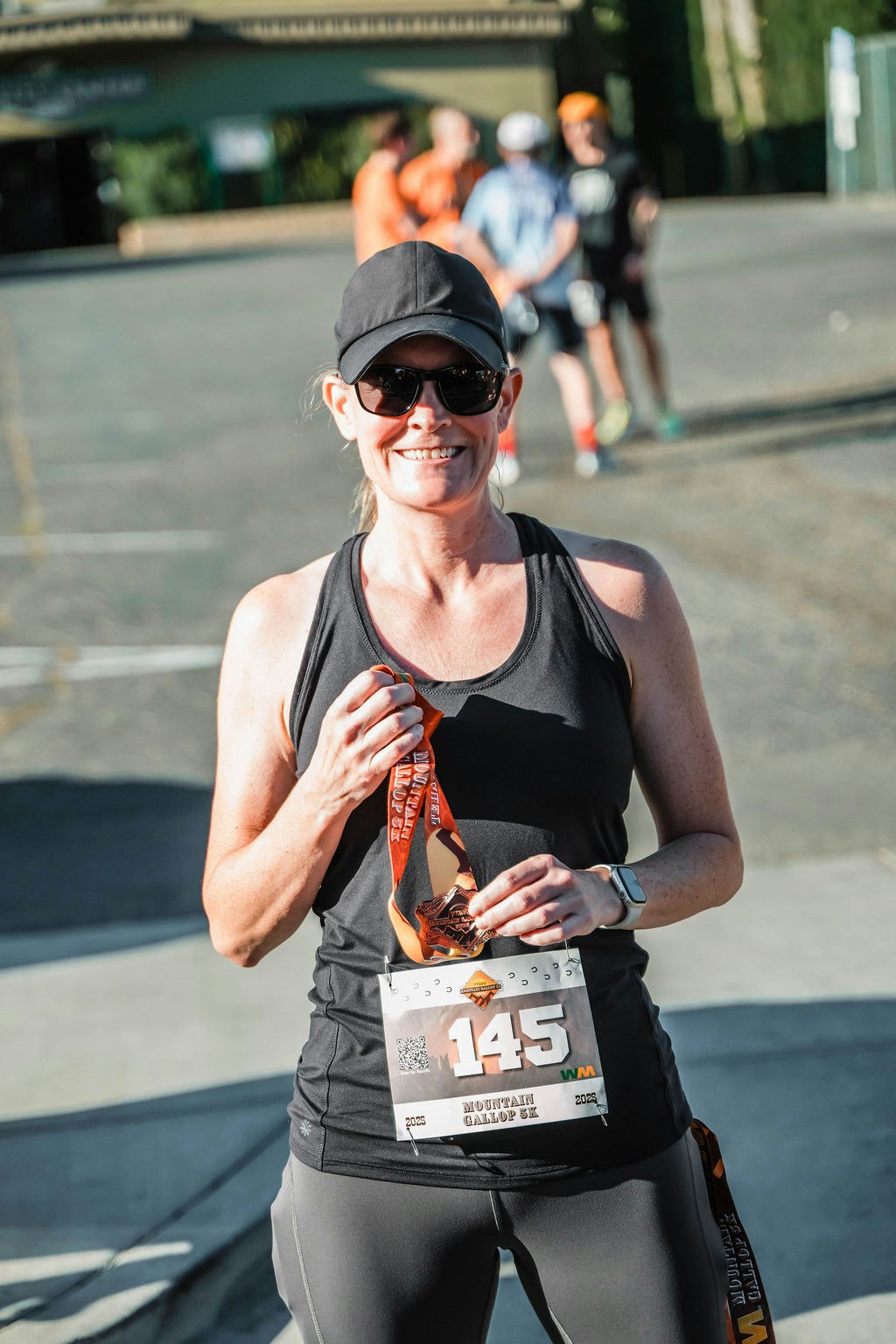 Smiling runner with a medal and race bib, wearing a cap and sunglasses, standing outdoors.