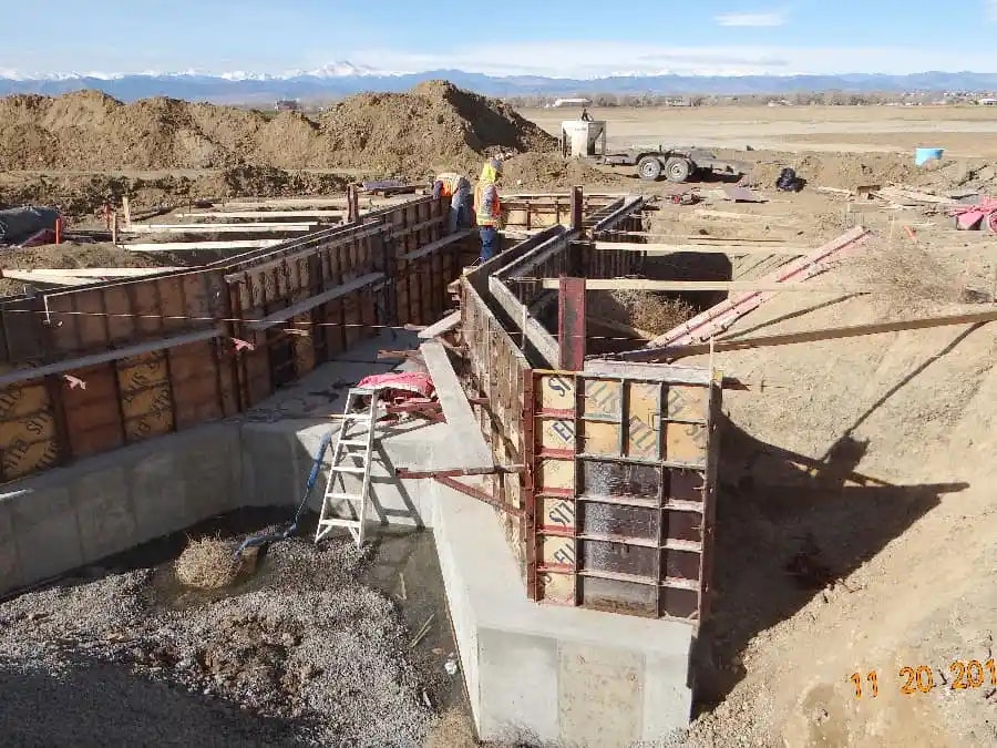 Construction site with workers and concrete forms, surrounded by piles of dirt and rural landscape.