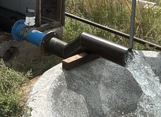 A metal pipe system discharging water into a stone basin, with visible vegetation in the background.