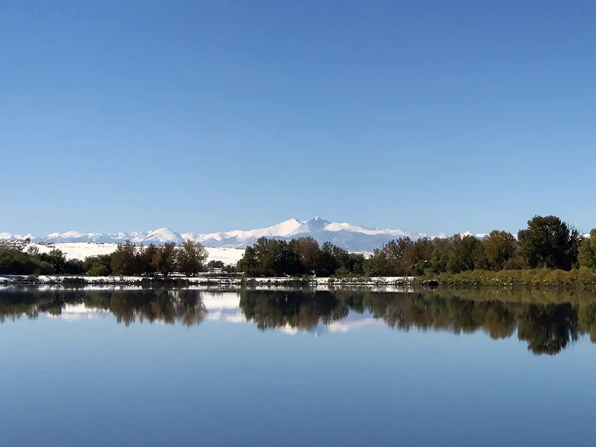 A calm lake reflecting trees and snow-capped mountains under a clear blue sky.