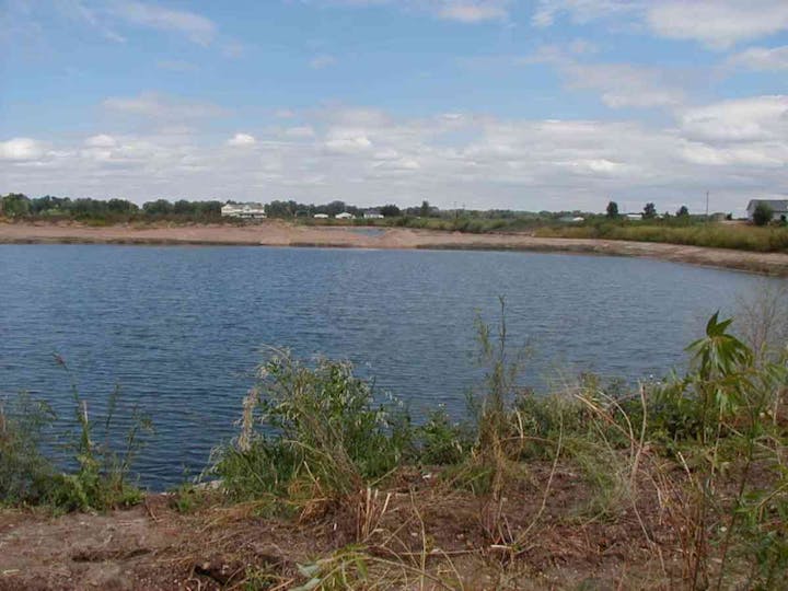 A small pond with surrounding vegetation and a distant horizon under a partly cloudy sky.