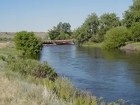 A calm river with green trees on both sides and a small bridge in the background.